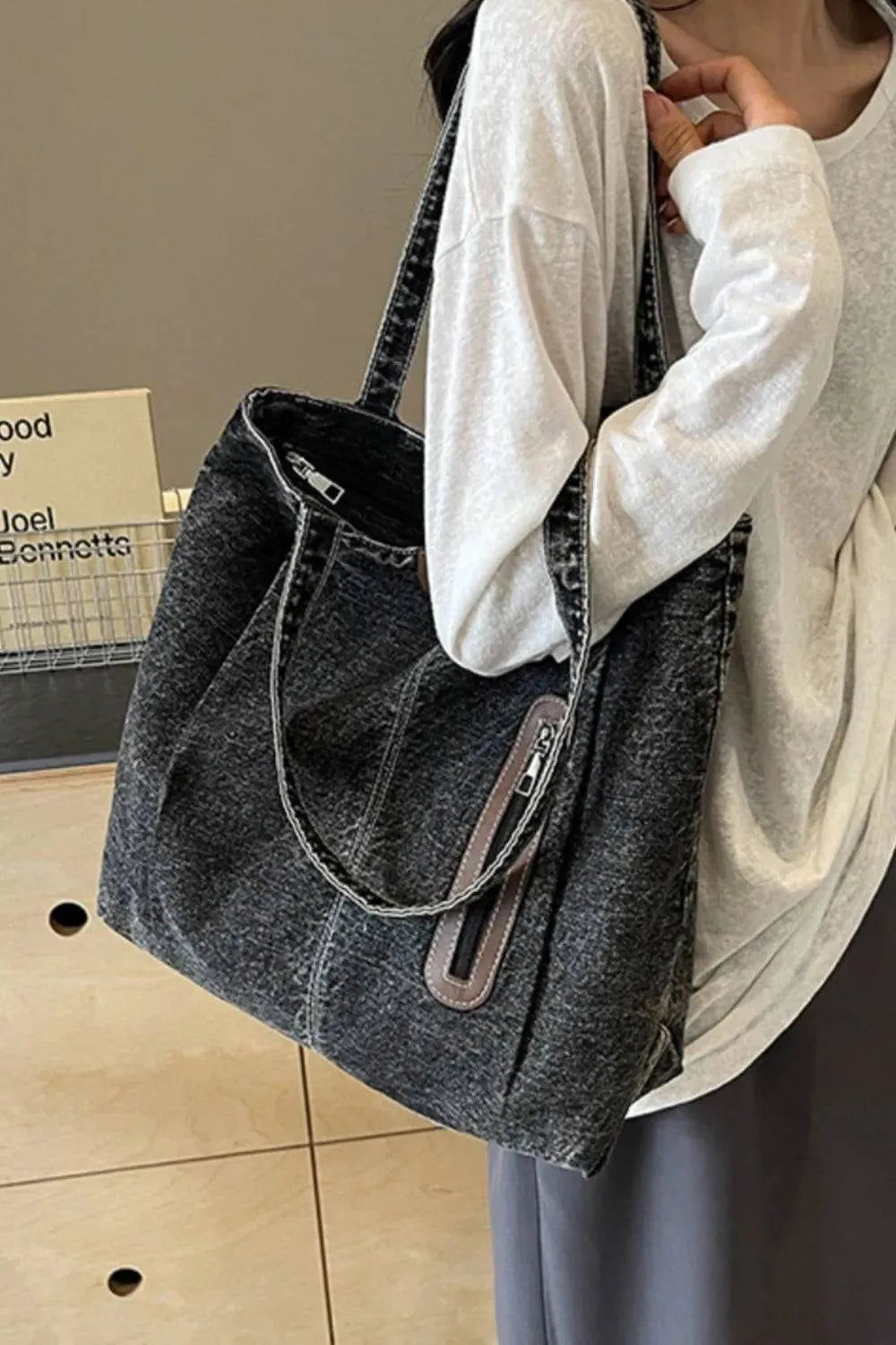 Eine Frau in einem lockeren weißen Langarmshirt und einem grauen Rock hält die flyliving Handtasche „Blue Jeans“, Größe M, mit Reißverschlusstasche und dezentem Logo. Im Hintergrund ist ein Holztisch mit Büchern zu sehen.