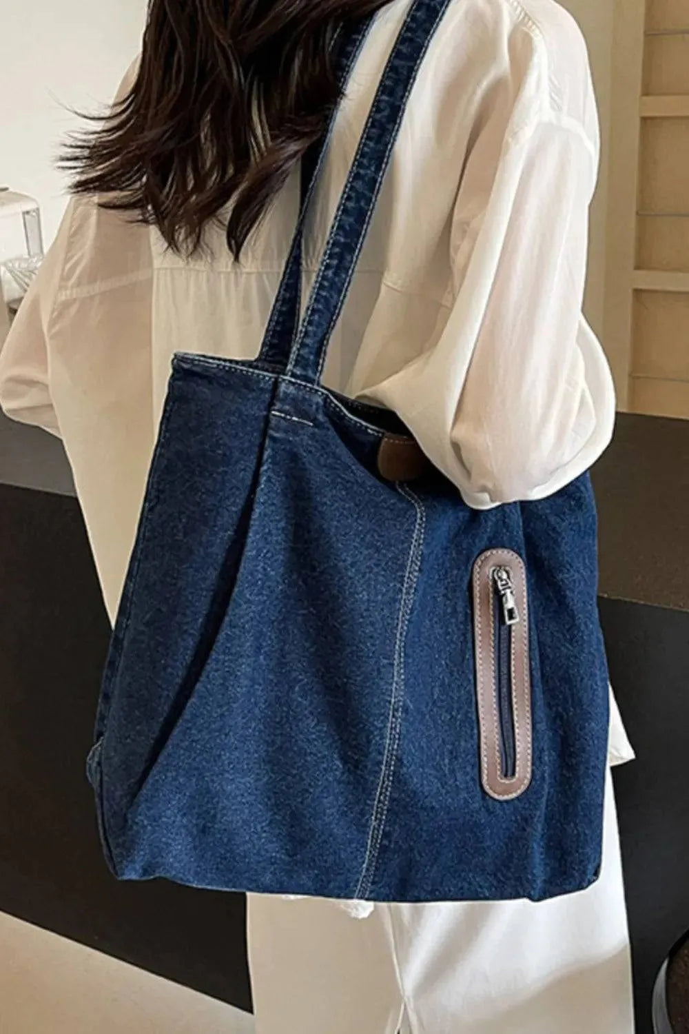Eine Person im weißen Hemd trägt die flyliving Handtasche „Blue Jeans“, medium für Frauen, mit Reißverschlusstasche vorne, breiten Riemen und dezentem Markenlogo, über der Schulter.