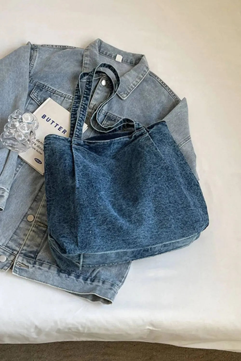 Die Handtasche „Blue Jeans“, mittelgroß, für Frauen, von flyliving liegt auf einer hellblauen Jeansjacke auf einer weißen Fläche, in deren Nähe teilweise eine durchsichtige Haarspange und ein mit BUTTER beschriftetes Papier zu sehen sind.