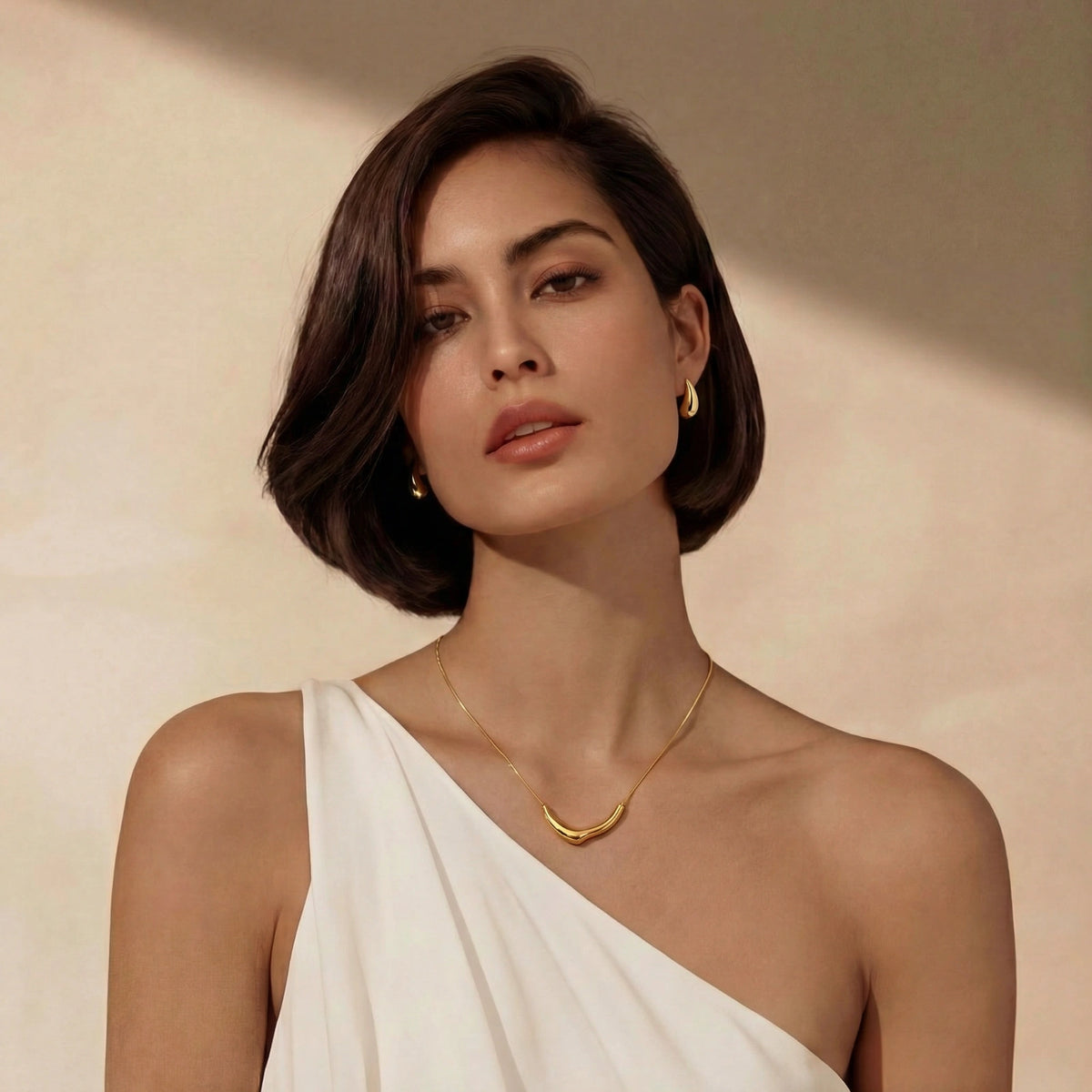 Frau mit dunklem Bob trägt goldene Tropfen Ohrstecker und Goldkette zu weißem One-Shoulder-Oberteil
