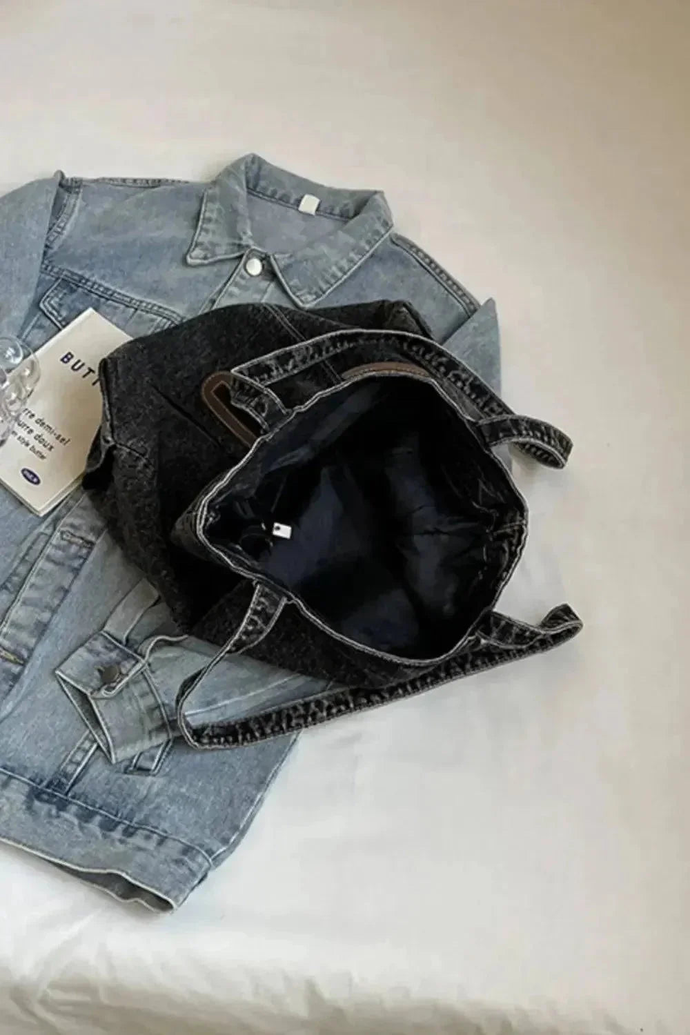 "Eine flyliving Handtasche „Blue Jeans“ (medium, für Frauen), eine robuste Denimjacke und ein gefaltetes Blatt Papier liegen auf weißem Untergrund; Die Handtasche ruht oben auf der Jacke."
