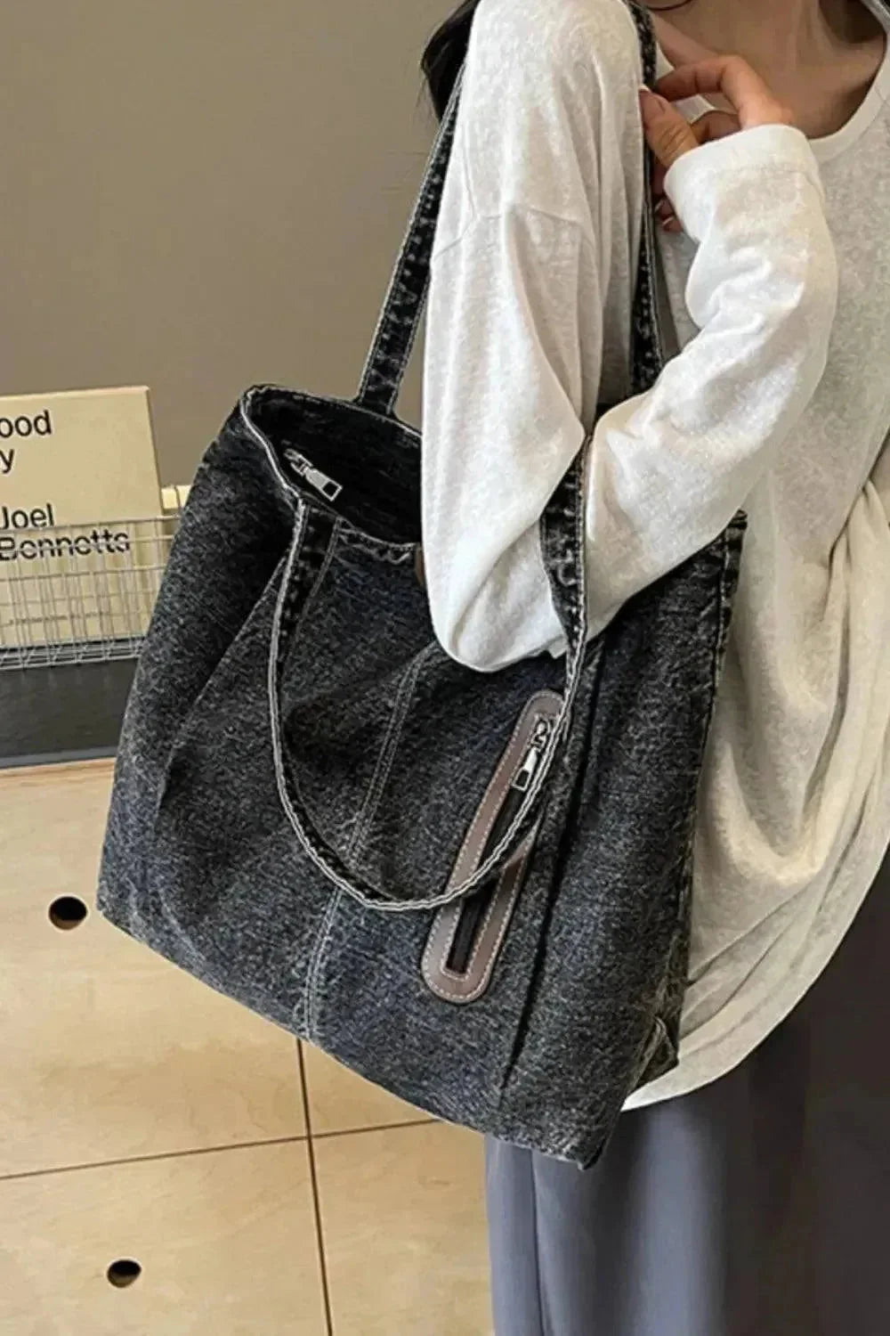 "Eine Frau in einem lockeren weißen Langarmshirt und einem grauen Rock hält die flyliving Handtasche „Blue Jeans“, Größe M, mit Reißverschlusstasche und dezentem Logo. Im Hintergrund ist ein Holztisch mit Büchern zu sehen."