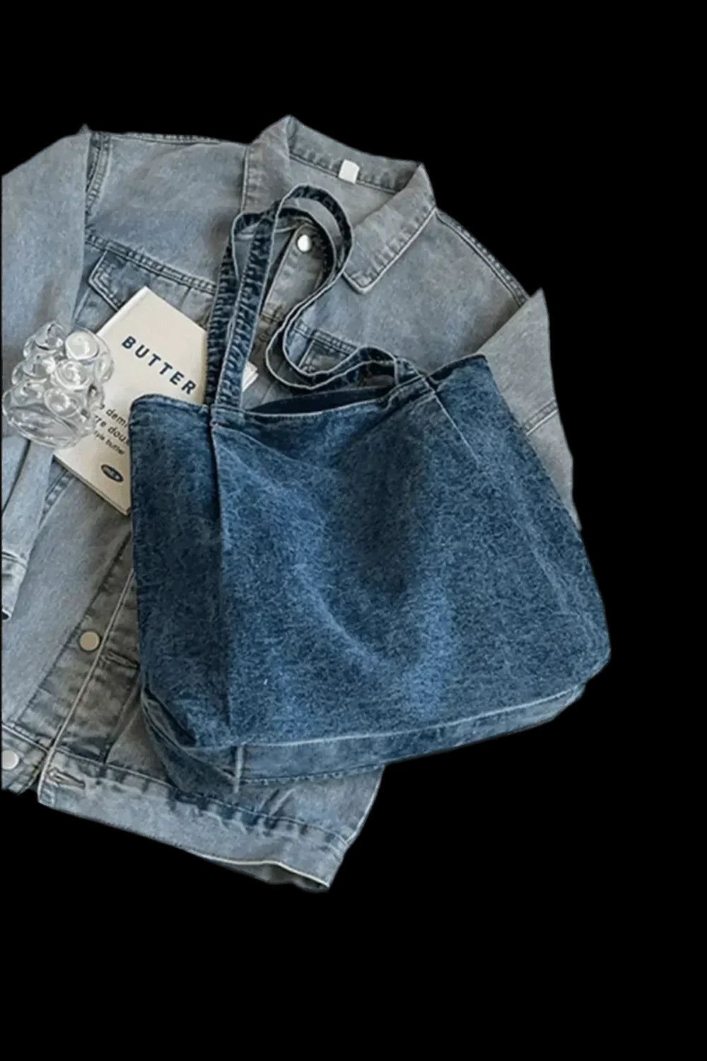 "Eine Handtasche „Blue Jeans“, Größe M für Damen, von flyliving liegt auf einer flach hingelegten Jeansjacke, daneben ein Heft mit der Aufschrift „BUTTER“ und ein durchsichtiger Plastikgegenstand."
