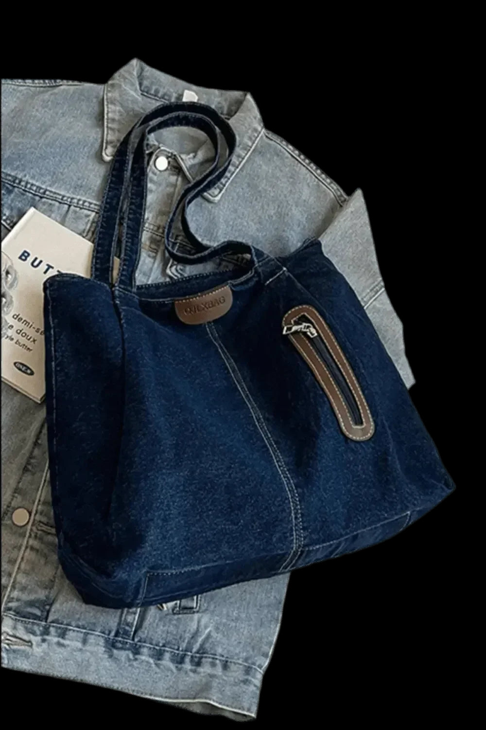 "Die flyliving Handtasche „Blue Jeans“, medium, für Frauen, ist eine dunkelblaue Denim-Tote mit braunen Akzenten, Frontreißverschlussfach, zwei Henkeln und dezentem Logo – platziert auf einer hellblauen Jeansjacke."