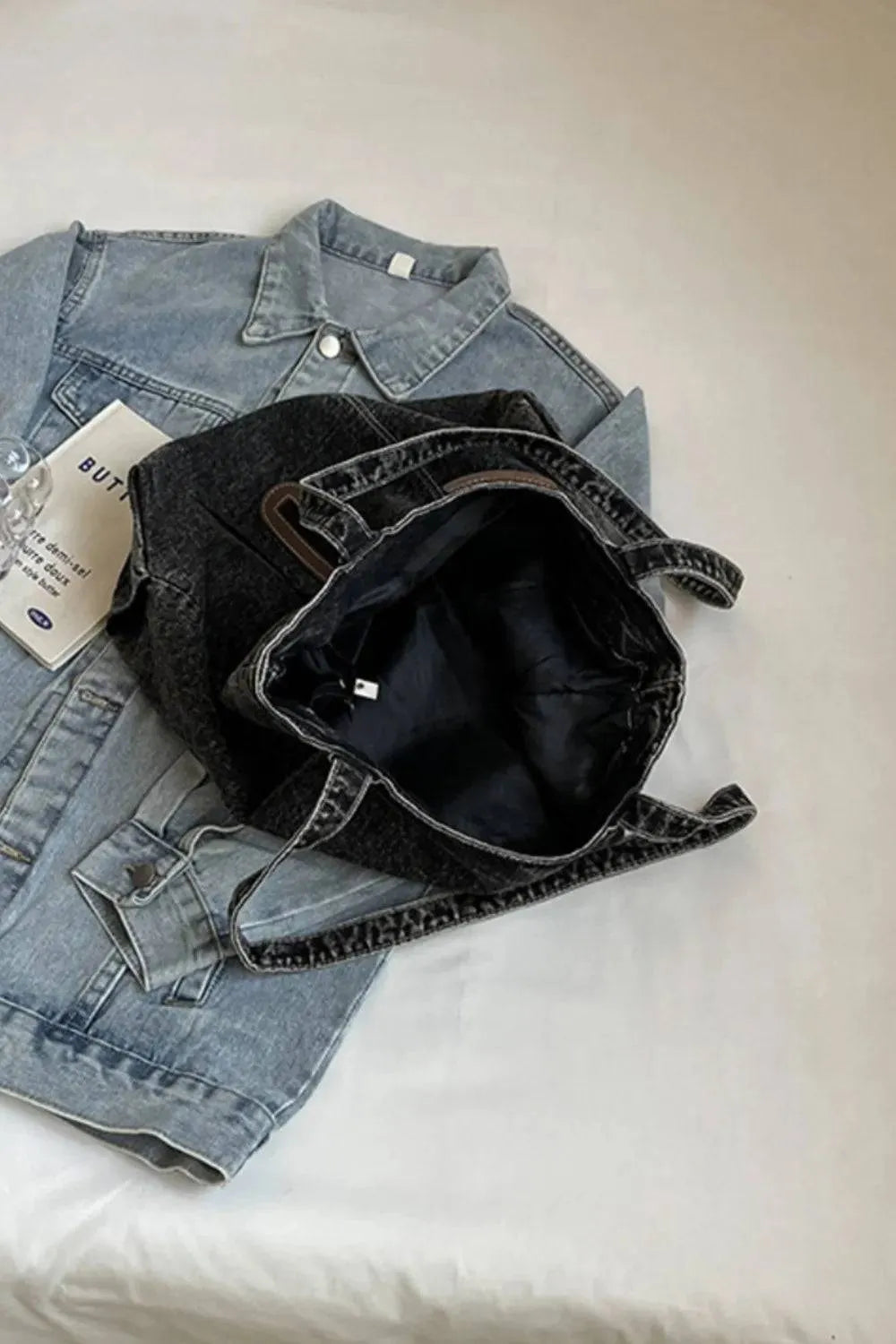 Eine flyliving Handtasche „Blue Jeans“ (medium, für Frauen), eine robuste Denimjacke und ein gefaltetes Blatt Papier liegen auf weißem Untergrund; Die Handtasche ruht oben auf der Jacke.