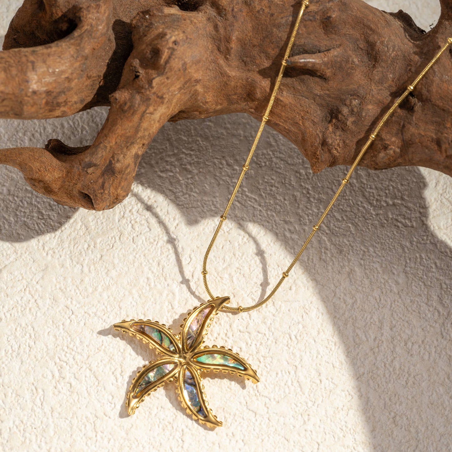 Gold-toned stainless steel starfish necklace with iridescent mother-of-pearl inlay and beaded chain by flyliving, on a textured surface with driftwood.