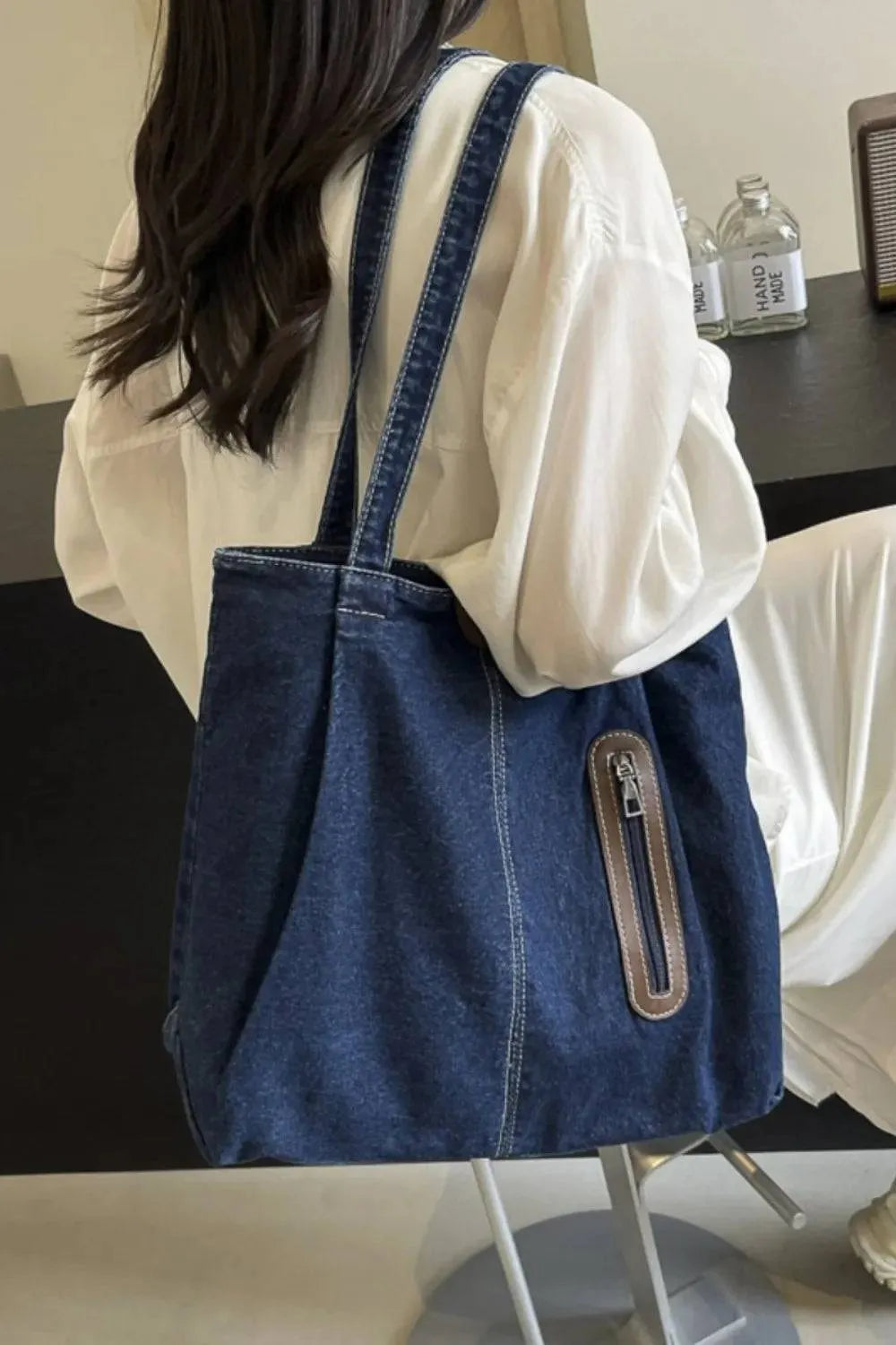 Eine Person mit langen dunklen Haaren in einem weißen Outfit sitzt auf einem Stuhl und trägt die flyliving Handtasche „Blue Jeans“, medium, für Frauen. Flaschen und ein Lautsprecher stehen auf dem Tisch dahinter.