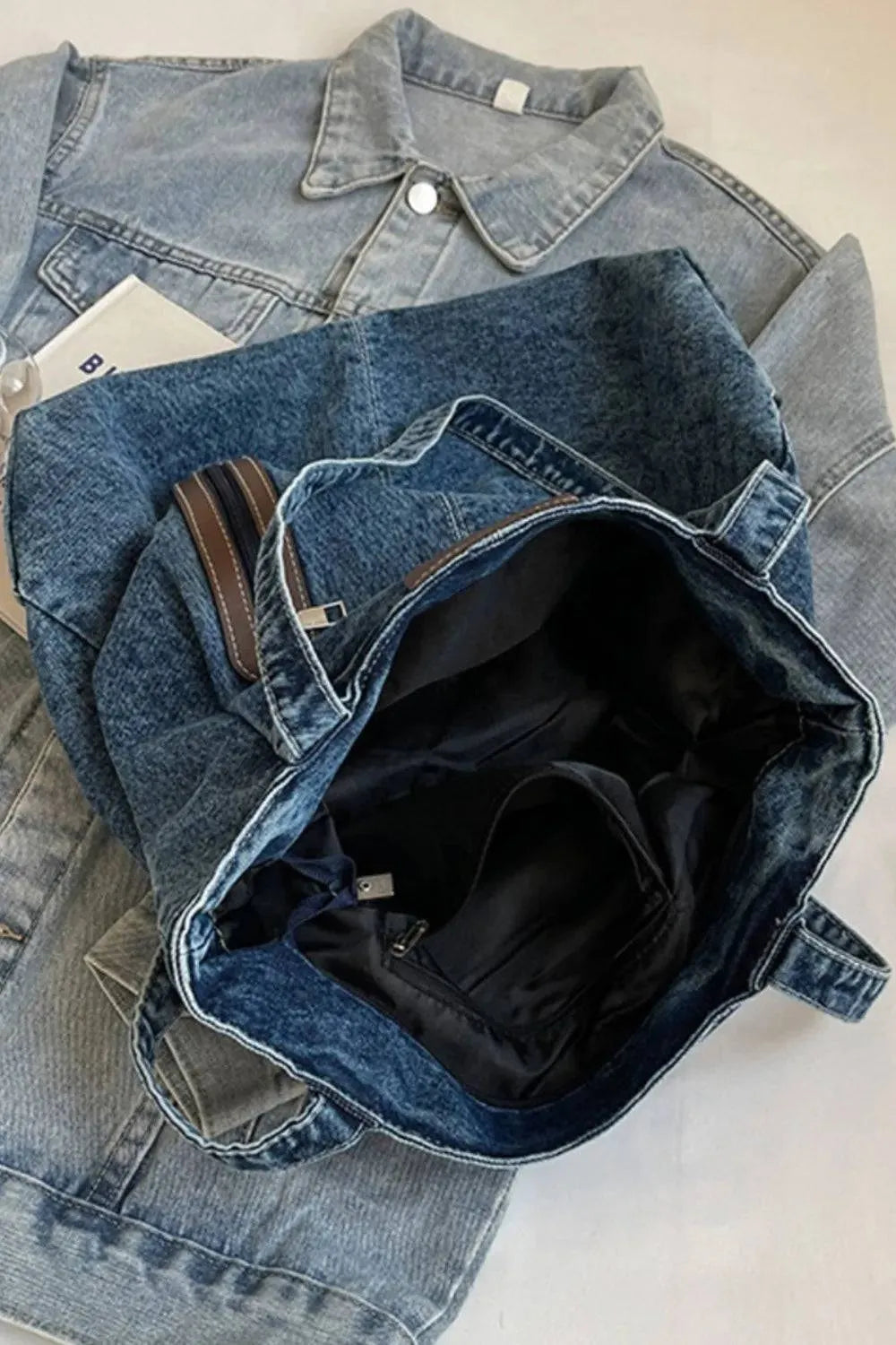 Die flyliving Handtasche „Blue Jeans“, mittel, für Frauen aus robustem Denim mit schwarzem Innenfutter liegt auf einer hellblauen Jeansjacke. Ein Brand-Logo-Tag ist teilweise sichtbar. Die Tasche hat kurze Henkel und wirkt leer.