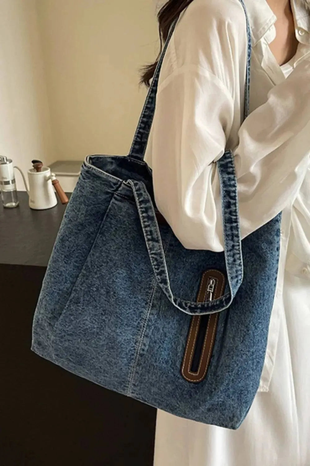 Eine Frau im weißen Hemd trägt die flyliving Handtasche „Blue Jeans“ (Medium) für Damen, die ein schickes Design mit braunen und silbernen Reißverschlussdetails aufweist. Drinnen steht sie an einem dunklen Tisch mit Teekanne und Tasse und präsentiert das dezente Markenlogo.