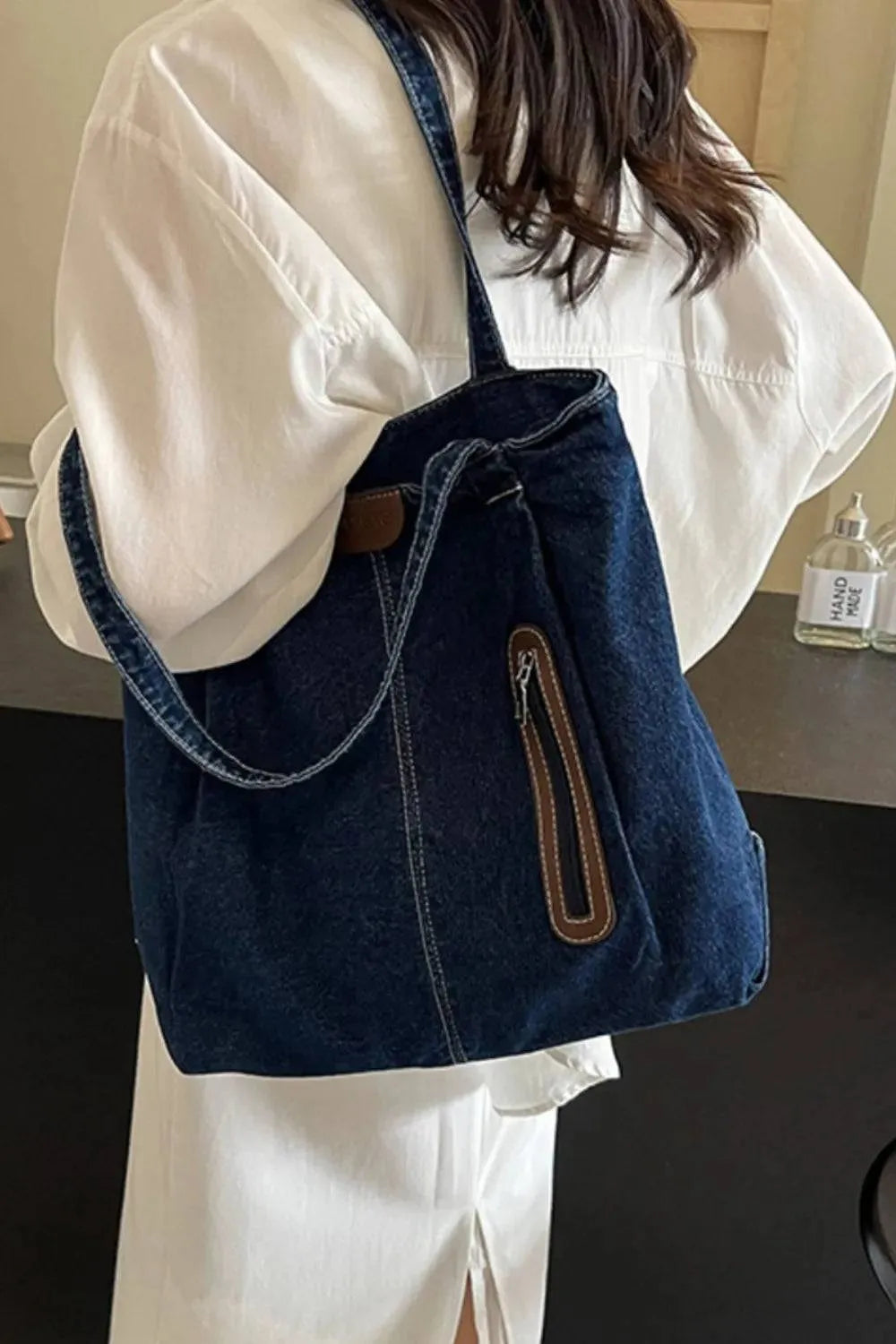 Eine Person im weißen Outfit trägt die mittelgroße Handtasche „Blue Jeans“ von flyliving mit braunem Reißverschluss und dezentem Markenlogo über der Schulter. Ihre langen Haare sind teilweise sichtbar, da sie im Innenbereich steht.