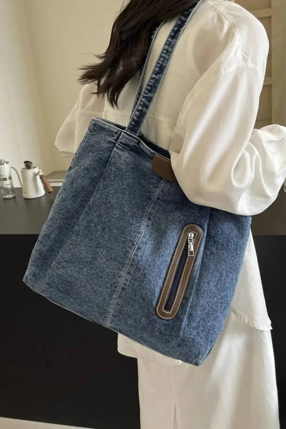 Eine Person in einem weißen Langarmkleid trägt die mittelgroße Damenhandtasche „Blue Jeans“ von flyliving mit einer Reißverschlusstasche vorne und einem braunen Etikett mit dem Markenlogo, während sie in einem Innenbereich vor einem schwarz-beigen Hintergrund steht.