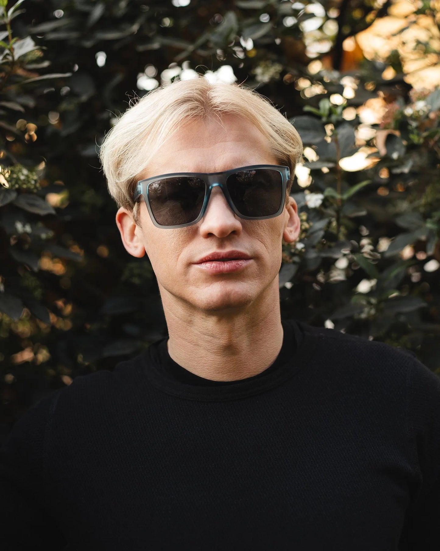 Eine Person mit kurzen, hellblonden Haaren, die INDICOM BRANDS SL Sonnenbrille Breezy Blue Hanukeii mit UV400 Schutz und ein schwarzes Oberteil trägt, steht vor grünem Laub und schaut direkt in die Kamera.