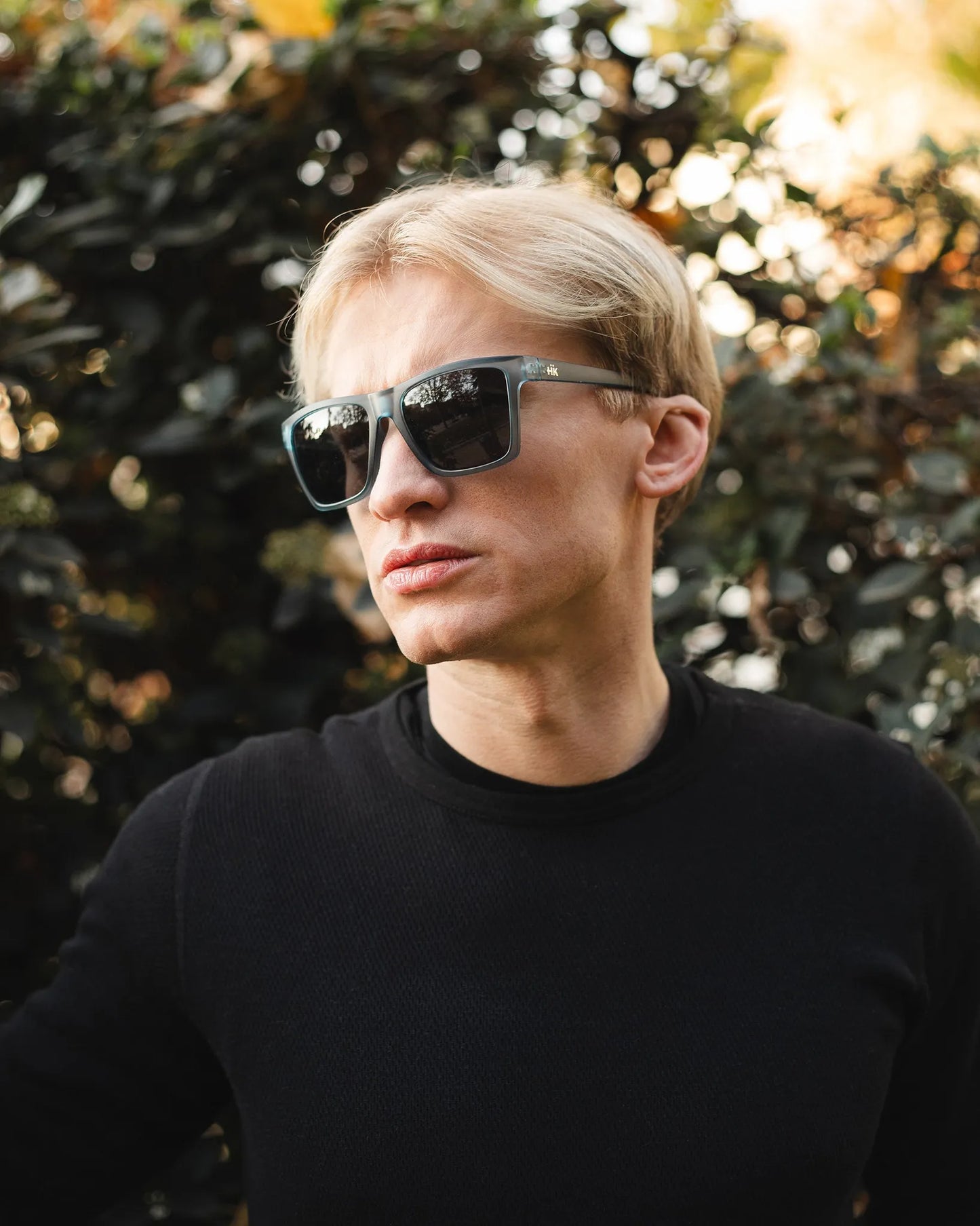 Eine Person mit kurzen blonden Haaren, bekleidet mit einem schwarzen langärmeligen Hemd, steht im Freien vor grünem Laub und trägt die Sonnenbrille Breezy Blue Hanukeii von INDICOM BRANDS SL mit Polycarbonat-Gläsern und UV400 Schutz.