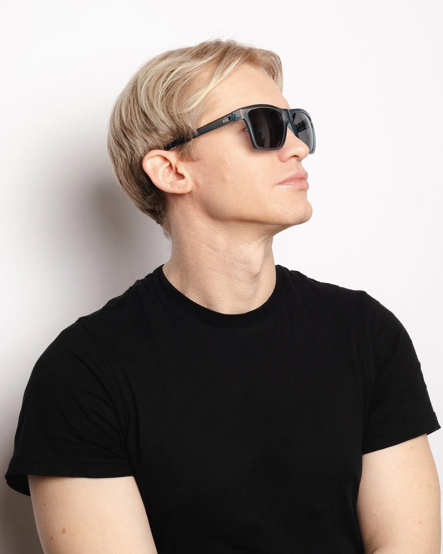 Eine Person mit kurzen blonden Haaren, die INDICOM BRANDS SL Sonnenbrille Breezy Blue Hanukeii mit UV400 Schutz und ein schwarzes T-Shirt trägt, schaut zur Seite vor einem schlichten weißen Hintergrund.