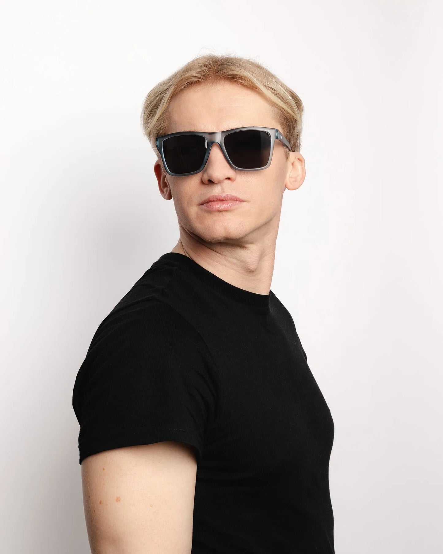 Ein Mann mit kurzen blonden Haaren, der ein schwarzes T-Shirt und INDICOM BRANDS SL Sonnenbrille Breezy Blue Hanukeii mit UV400 Schutz trägt, steht vor einem schlichten weißen Hintergrund und schaut zur Seite.