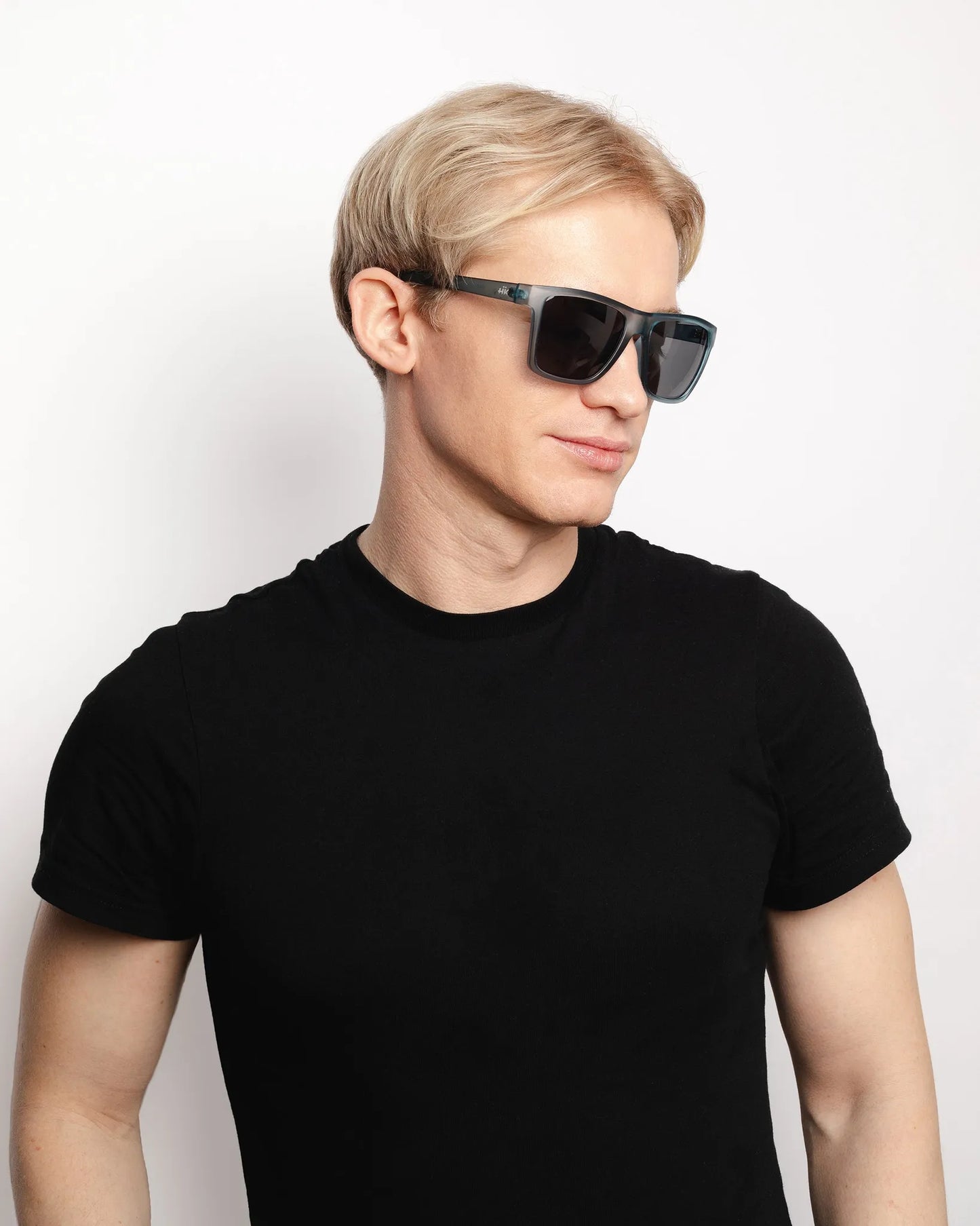 Ein Mann mit kurzen blonden Haaren trägt die Sonnenbrille Breezy Blue Hanukeii von INDICOM BRANDS SL, die mit Polycarbonat-Gläsern und UV400 Schutz ausgestattet ist. Er steht in einem schwarzen T-Shirt vor einem schlichten weißen Hintergrund und blickt mit einem neutralen Blick zur Seite.