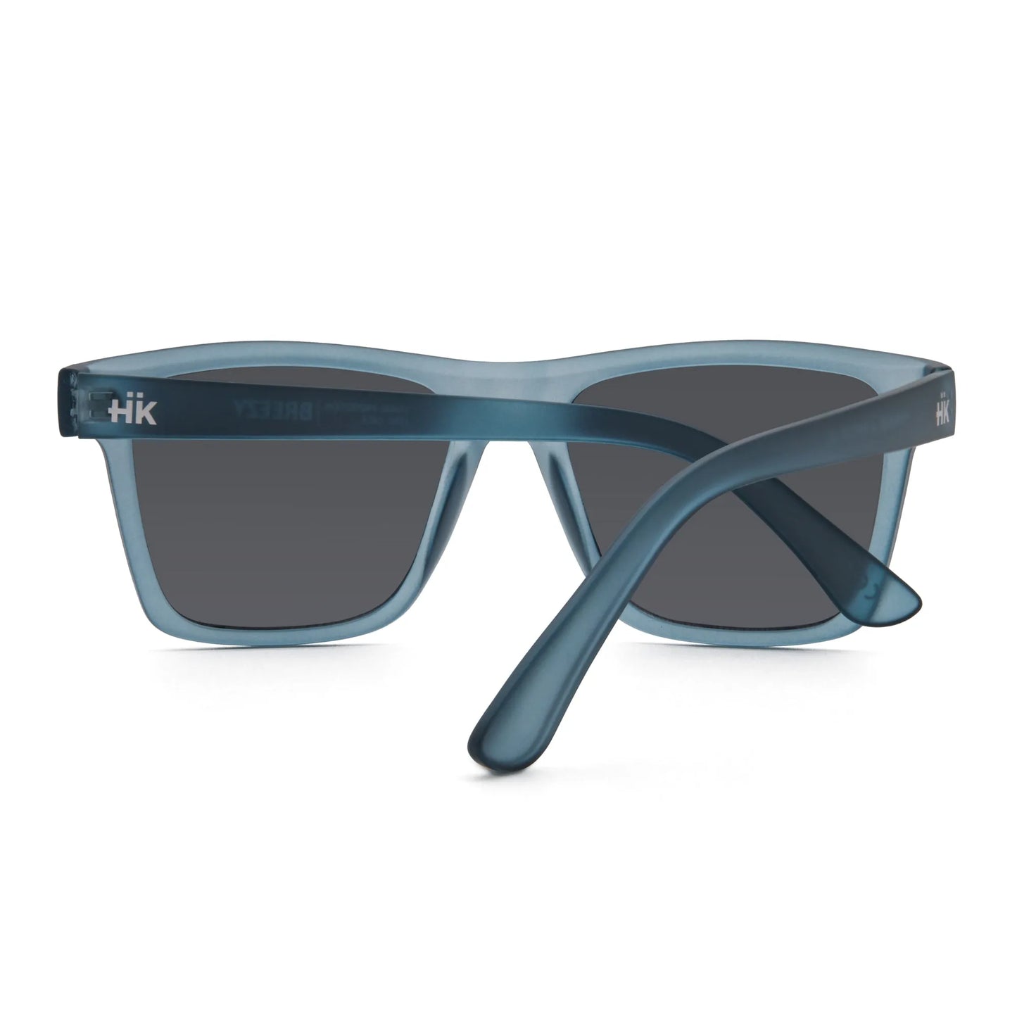 Die Sonnenbrille Breezy Blue Hanukeii von INDICOM BRANDS SL hat einen blauen, durchscheinenden Rahmen, dunkle Polycarbonat-Gläser mit UV400-Schutz und ein weißes "HK" auf dem linken Bügel, von hinten gesehen mit gekreuzten Armen.