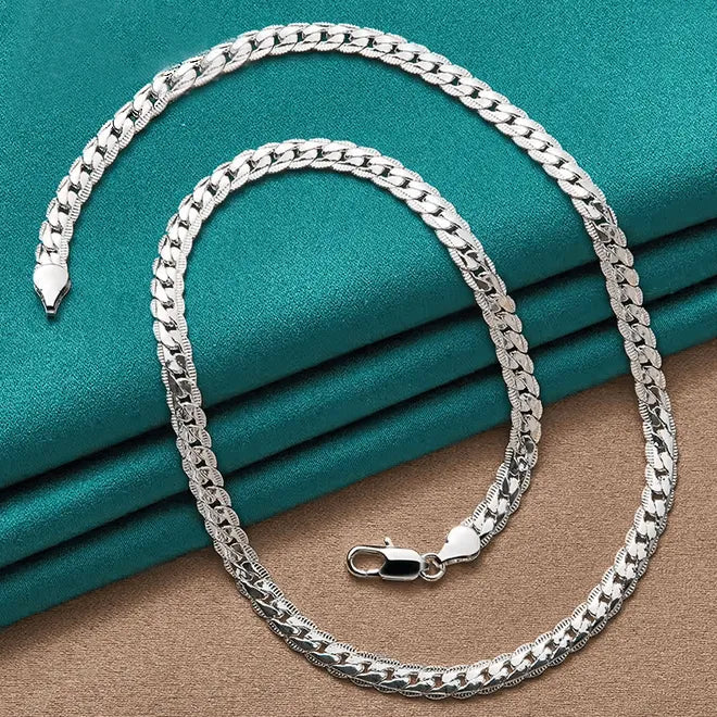 Die flyliving Halskette „Horst-Peter“, eine flache, glänzende 925er Sterlingsilber Gliederkette mit Karabinerverschluss, ist in einer lockeren Spirale auf türkis-beigem Stoff angeordnet.