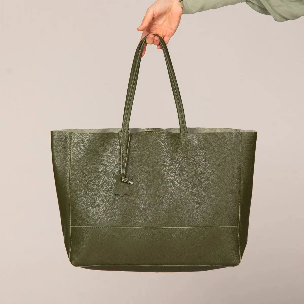 Eine Hand hält die grüne flyliving Umhängetasche "Susi" aus Leder, mit Doppelgriffen und einem kleinen Anhänger, vor einem schlichten beigen Hintergrund.