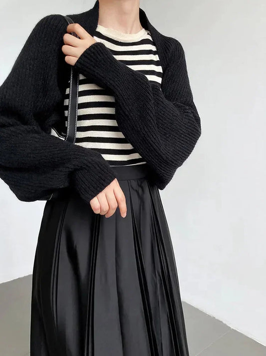 Eine Person trägt die marigoldshadows Touka Strick Shrug in schwarz über einem schwarz-weiß gestreiften Hemd und einem schwarzen Faltenrock und setzt damit moderne Damenmode vor einem schlichten hellen Hintergrund in Szene.