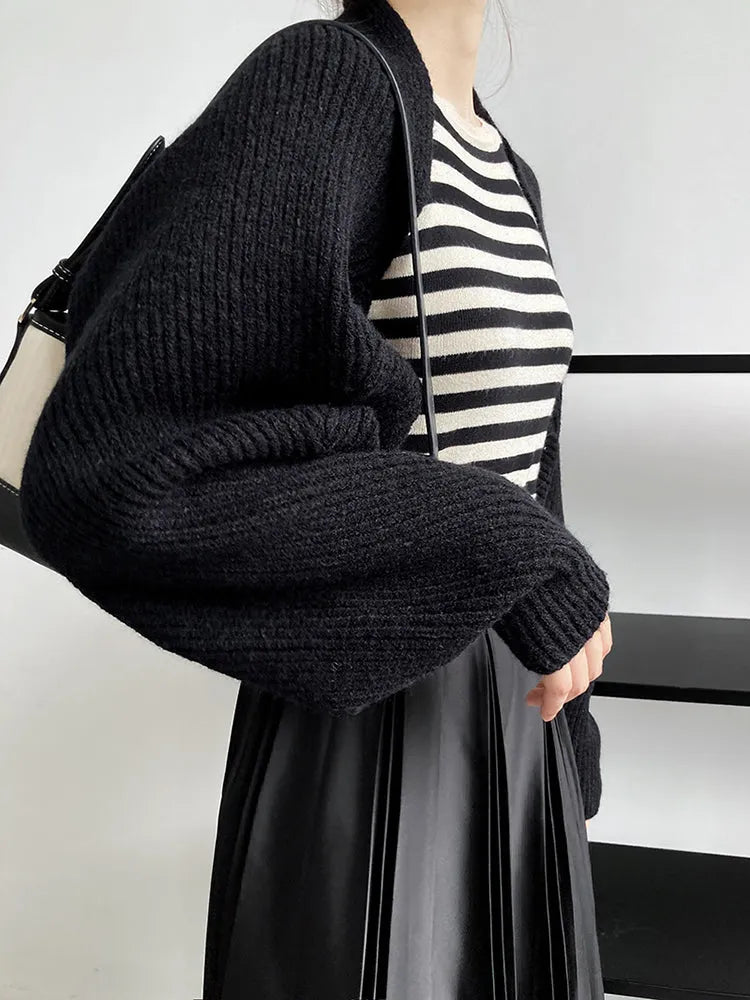 Eine Person trägt den Touka Strick Shrug in Schwarz von marigoldshadows über einem schwarz-weiß gestreiften Top und plissierten Rock, hält eine schwarz-weiße Tasche und steht in minimalistischer Umgebung.