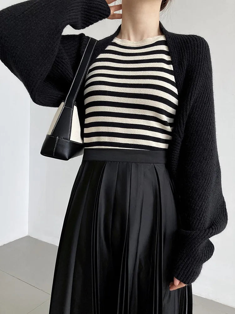 Eine Person trägt Damen Mode: schwarze Oberteile, darunter den Touka Strick Shrug in Schwarz von marigoldshadows, eine schwarz-weiß gestreifte Bluse, einen plissierten Rock und eine schwarze Umhängetasche. Der Kopf ist nicht sichtbar.