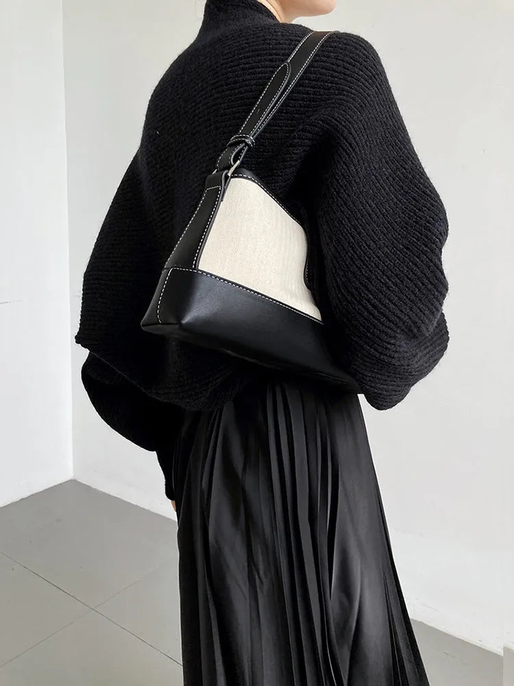 Eine Person, die den marigoldshadows Touka Strick Shrug in Schwarz trägt, gestylt mit einem Faltenrock und einer zweifarbigen Umhängetasche, steht vor einer schlichten, hellen Wand - ein schicker Damen Mode-Layering Inspo. Gesicht nicht sichtbar.
