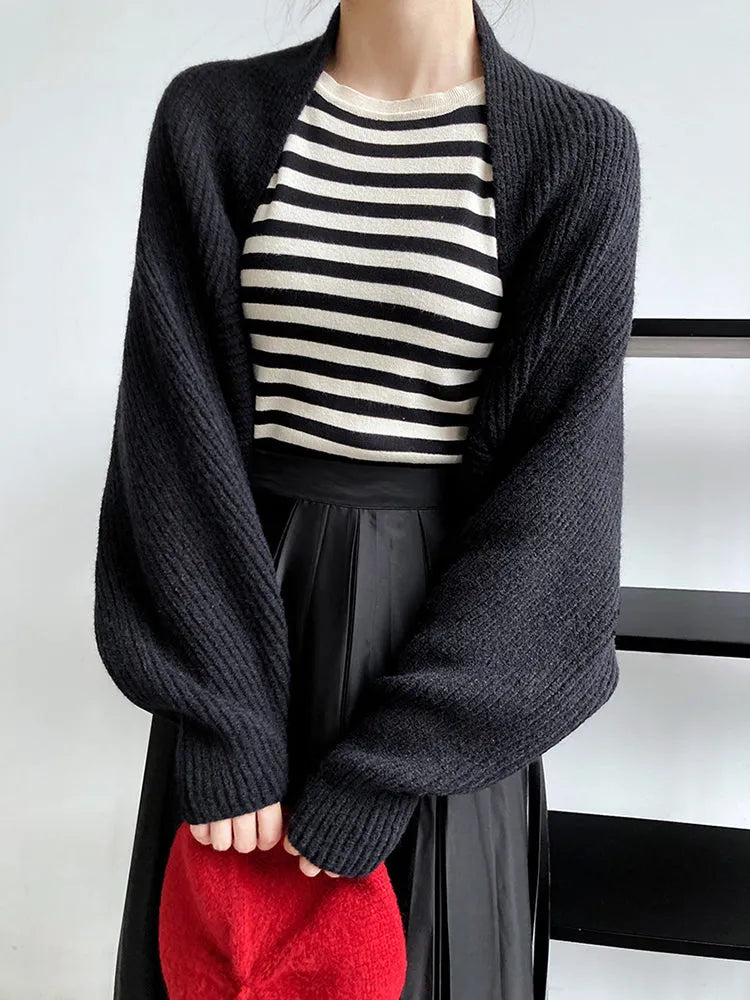 Eine Person trägt die Marigoldshadows Touka Strick Shrug in Schwarz über einem schwarz-weiß gestreiften Hemd und einem schwarzen Rock und hält ein rotes Wuschelteil. Der Hintergrund ist einfach mit einem schwarzen Regal; das Gesicht der Person ist nicht zu sehen.