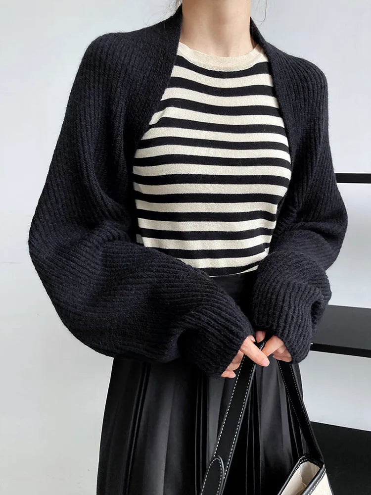 Eine Frau modelliert Damen Mode, trägt ein schwarz-weiß gestreiftes Oberteil, die marigoldshadows Touka Strick Shrug in schwarz und einen schwarzen Rock, während sie eine passende Handtasche hält. Nur ihr Oberkörper ist vor einem einfarbigen Hintergrund zu sehen.