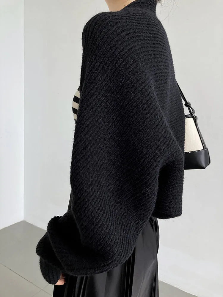 Eine Person trägt die marigoldshadows Touka Strick Shrug in schwarz über einem schwarz-weiß gestreiften Top, mit einem schwarzen Faltenrock und einer kleinen Umhängetasche. Dieser schicke Damen Mode Look ist drinnen vor einer schlichten Wand fotografiert.