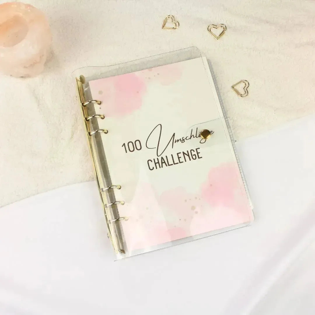 Der BeginnerBudget 100 Umschläge Sparchallenge-Ordner liegt auf einer weißen und cremefarbenen Oberfläche und wird durch goldene Herz-Büroklammern, beschriftete Umschläge und einen rosa Kerzenhalter akzentuiert - perfekt für den Start Ihrer Sparchallenge.
