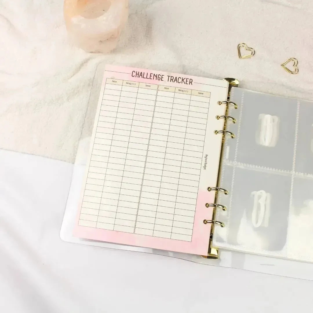 Ein aufgeschlagener BeginnerBudget 100 Umschläge Sparchallenge Planer zeigt eine Challenge Tracker Seite mit rosa Rand und leeren Spalten, die auf hellem Stoff neben einer Kerze und herzförmigen Büroklammern liegt - ideal für Ihre nächste Spar-Challenge.
