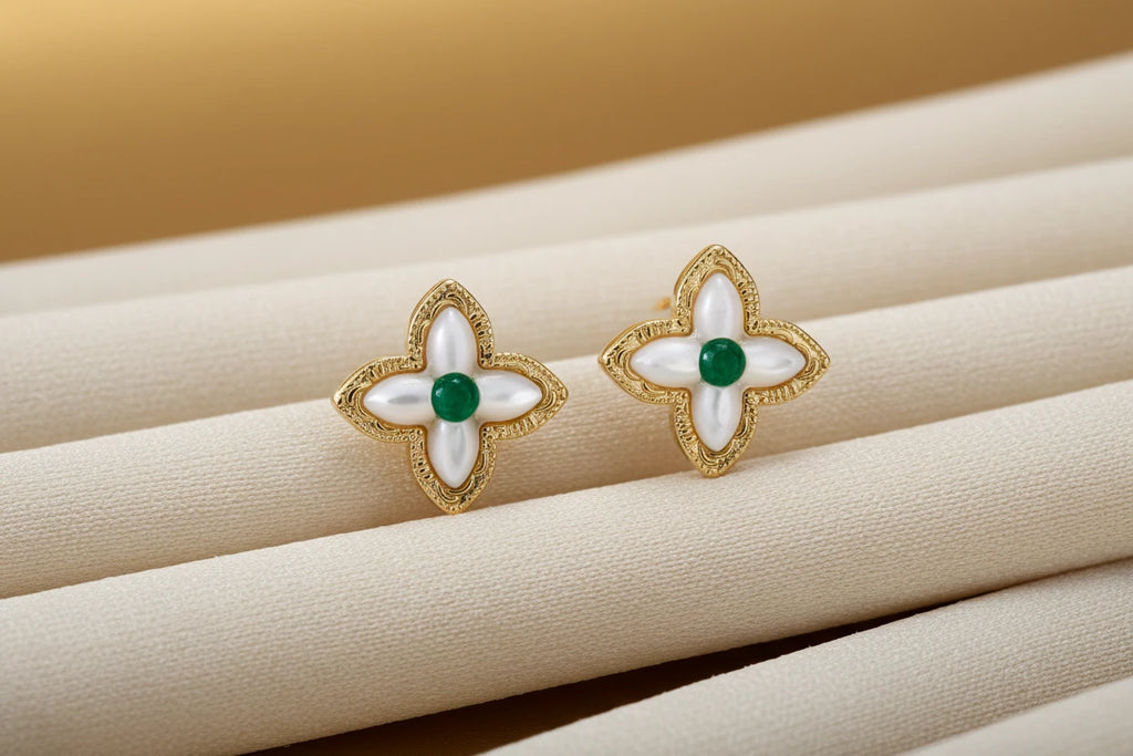 Vergoldete Glücksklee-Ohrringe von flyliving, mit grünen Akzenten und perlmuttfarbenen Details auf einem beigen Stoffhintergrund.
