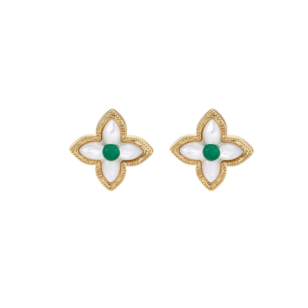 Gold quatrefoil stud earrings with white mother-of-pearl petals and emerald green gemstone center on beige background.