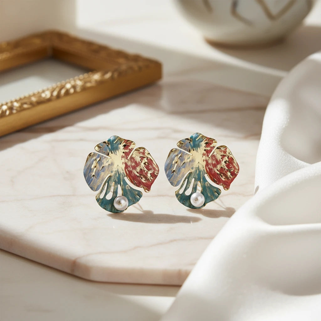 Vergoldete Monstera Muse Ohrringe von flyliving, mit Emaille-Details und Perlenakzenten, auf Marmorplatte arrangiert.