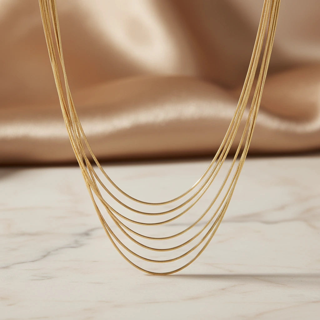 Mehrreihige goldfarbene Halskette von flyliving, filigran und glänzend, vor beigem Stoff und Marmorplatte.