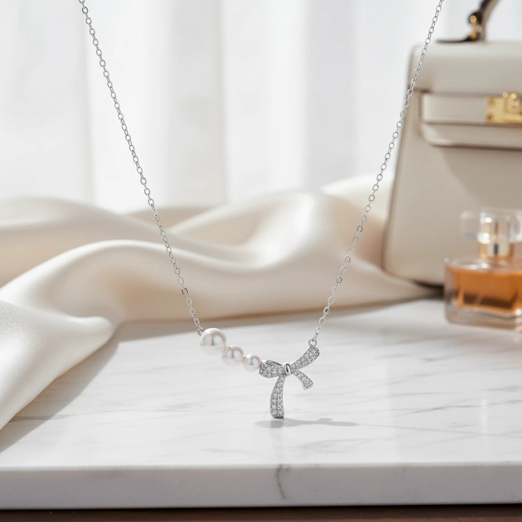 Versilberte Perlen-Schleifen-Halskette von flyliving, mit weißen Perlen und glitzerndem Schleifenanhänger, vor einem verschwommenen Hintergrund.
