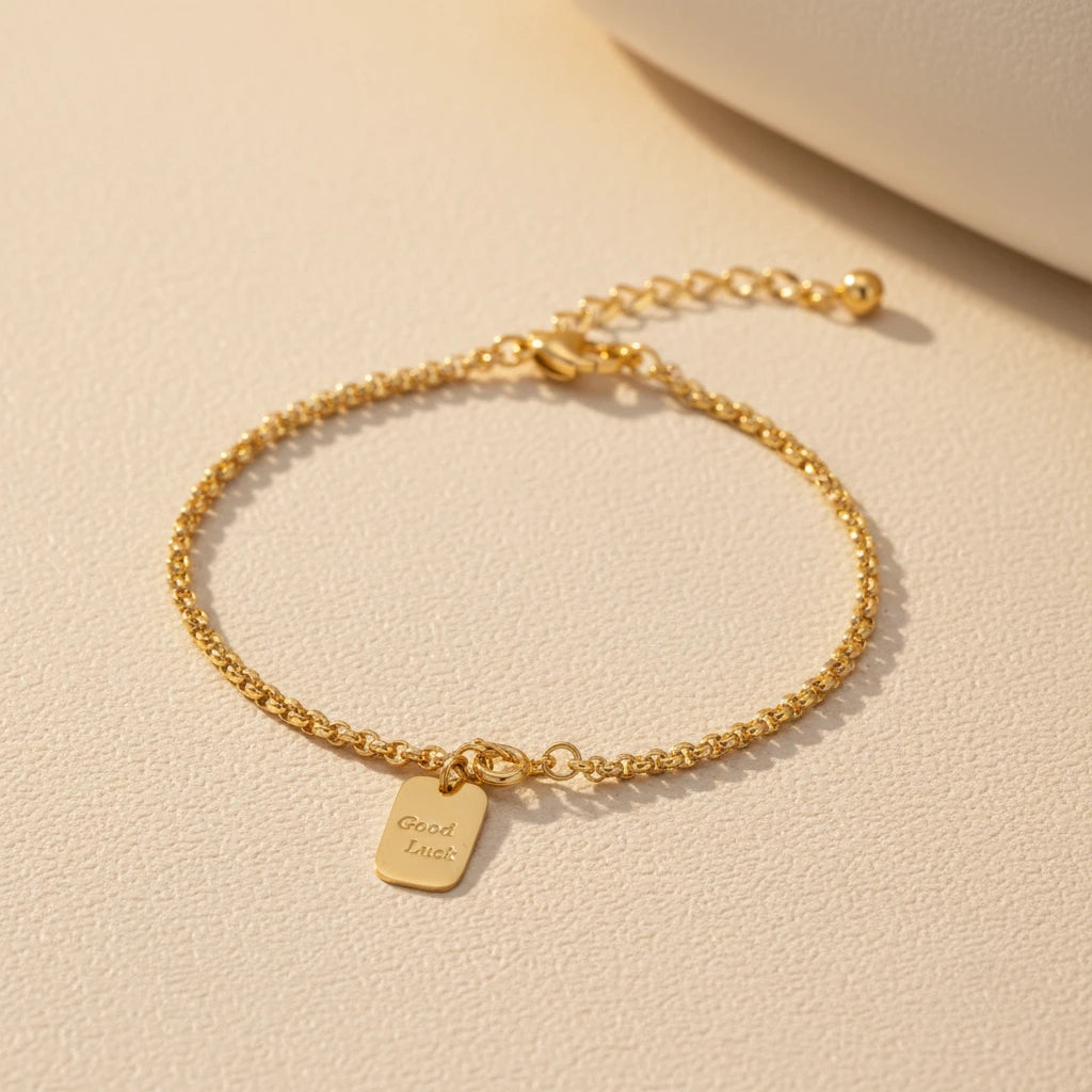 Vergoldetes Armband "Good Luck" von flyliving mit kleinem, rechteckigem Anhänger auf beigem Untergrund.