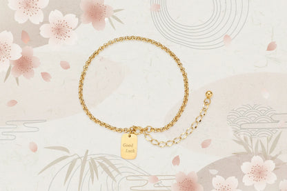 Vergoldetes "Good Luck" Armband von flyliving mit dekorativen Kirschblüten und Wellenmuster auf beigem Grund.