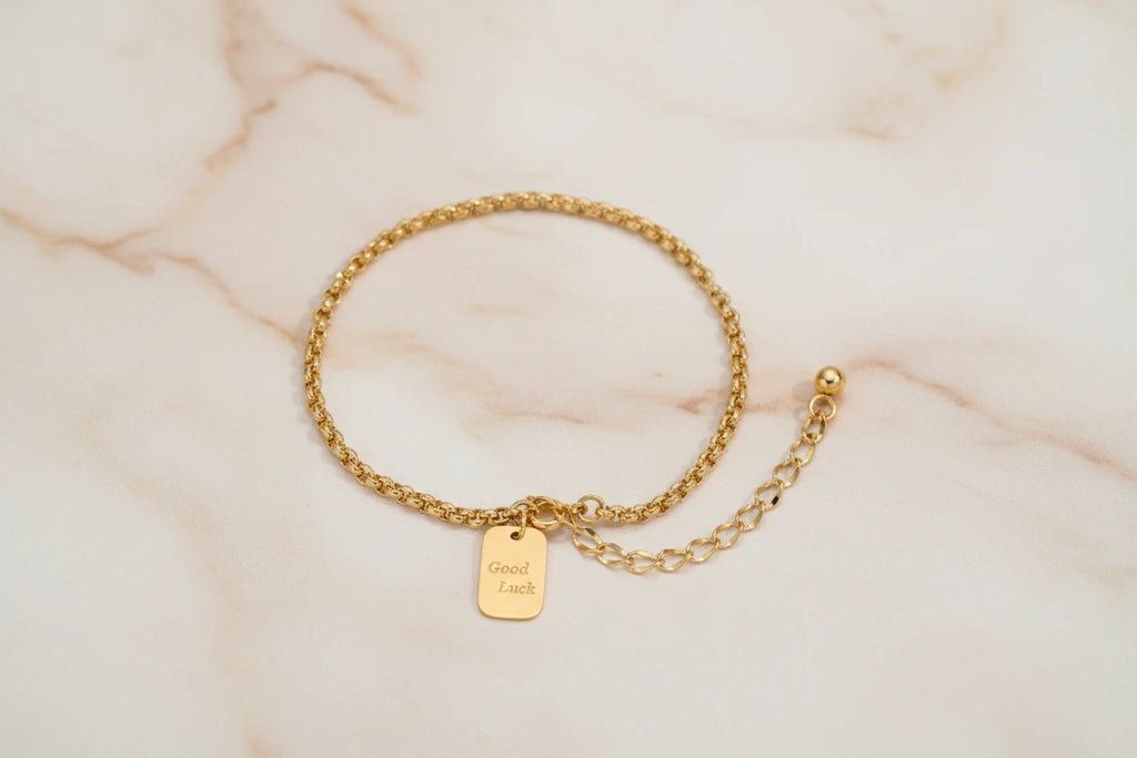Vergoldetes Good Luck Armband von flyliving, mit rechteckigem Anhänger und feingliedriger, verstellbarer Kette auf Marmor.
