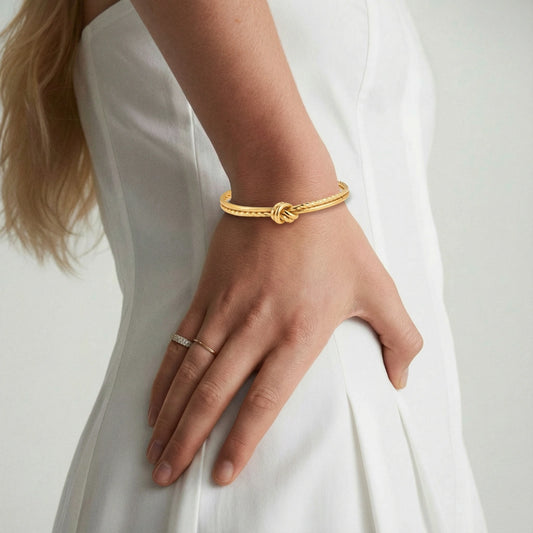 Goldenes Knotenarmband, vergoldet von flyliving, an einem weiblichen Handgelenk mit goldenen Ringen, vor einem weißen Kleid.