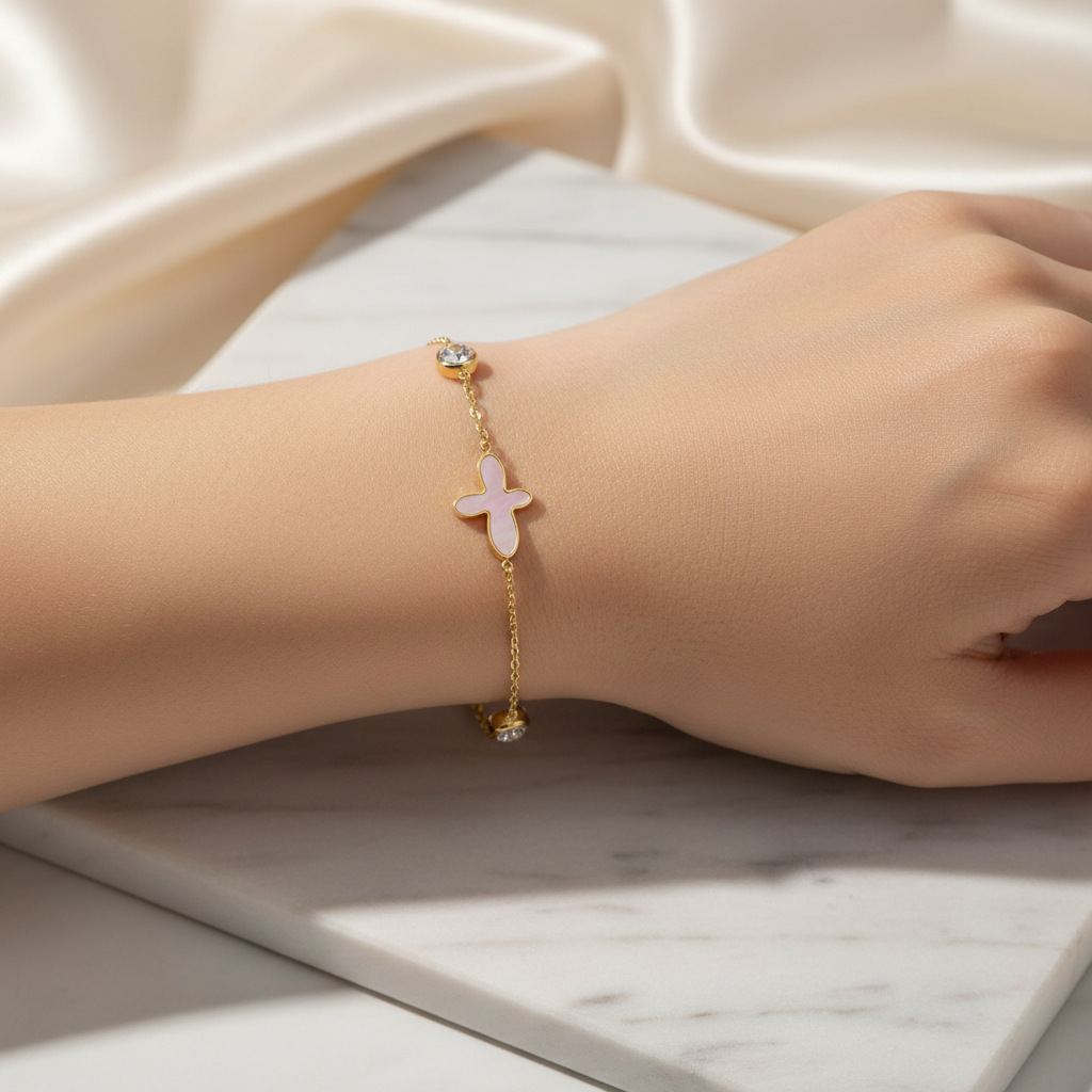 18K gold-plated bracelet with a pink mother-of-pearl cross charm and crystals by flyliving, worn on a wrist on marble with satin background.