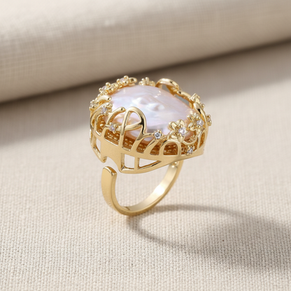 Goldfarbener Ring mit großer weißer Barockperle in durchbrochener Krone mit Blüten und Zirkonia auf beigem Stoff