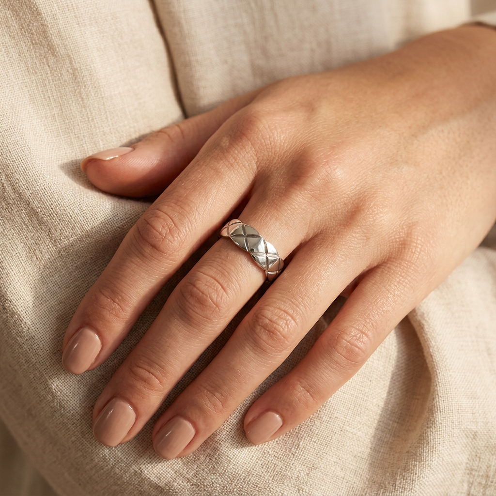 Silver-toned ring with a quilted pattern by flyliving, worn on a hand with nude nail polish against beige fabric.