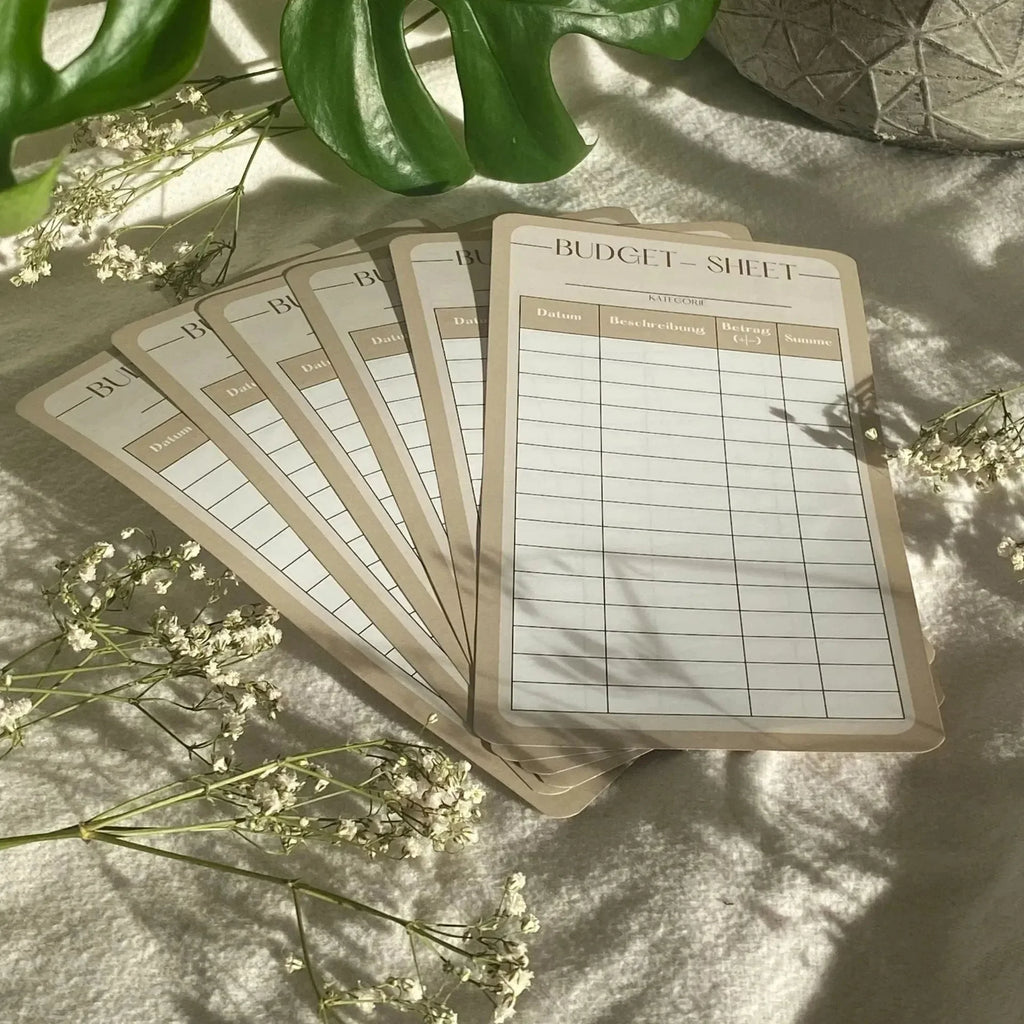 Zehn beigefarbene Budgetblätter für Anfänger (10er-Pack) liegen auf einer weichen Oberfläche mit weißen Blumen und grünen Blättern, die in Sonnenlicht getaucht sind - eine einladende Umgebung, um mit der Organisation Ihres Sparplans oder Haushaltsbuchs zu beginnen.