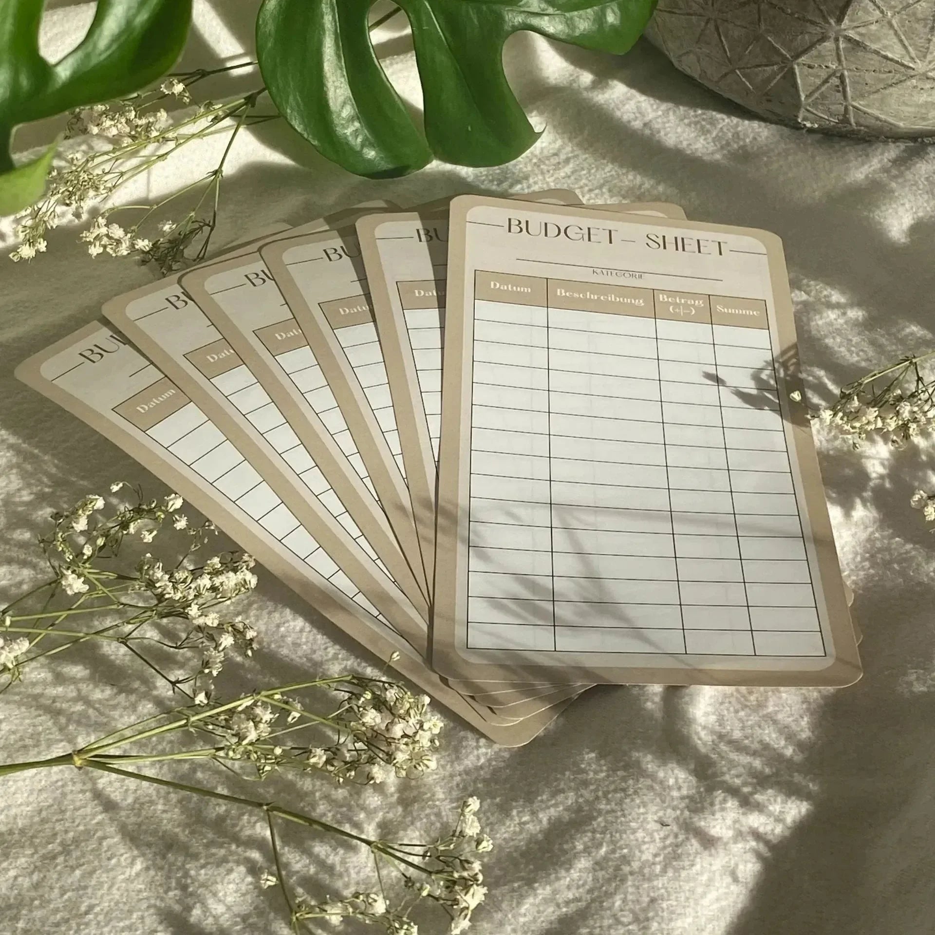 Zehn beigefarbene Budgetblätter für Anfänger (10er-Pack) liegen auf einer weichen Oberfläche mit weißen Blumen und grünen Blättern, die in Sonnenlicht getaucht sind - eine einladende Umgebung, um mit der Organisation Ihres Sparplans oder Haushaltsbuchs zu beginnen.