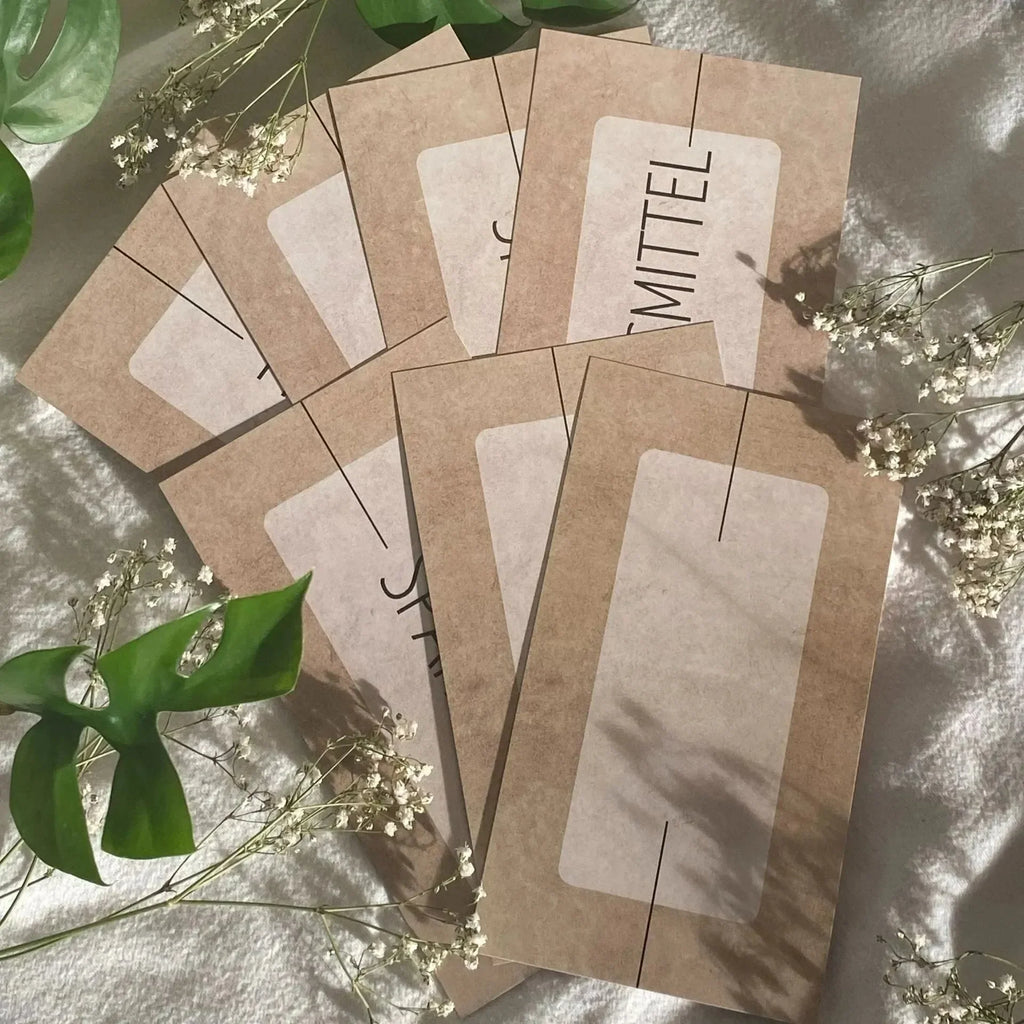 Sieben braune Umschläge mit weißen Etiketten aus der Grundausstattung Budget Planung von Budget Binder von BeginnerBudget sind auf einem hellen Hintergrund mit grünen Blättern und kleinen weißen Blumen aufgefächert - perfekt für die Umschlagmethode zum Budgetieren und Bargeldtracker.