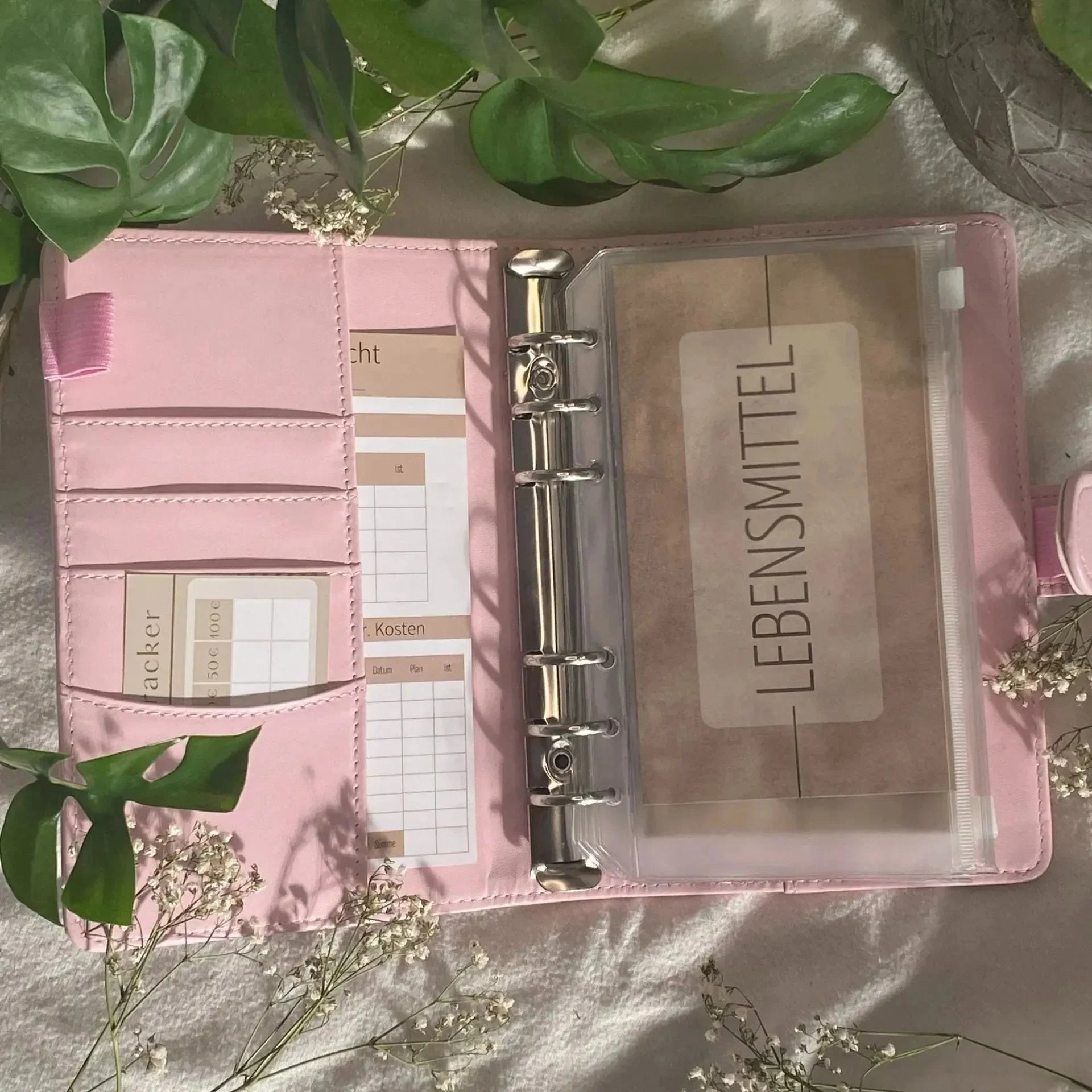 Die "Grundausstattung Budget Planung von Budget Binder" von BeginnerBudget, ein pinker Organizer mit beschrifteten Umschlägen und Budgetblättern, liegt auf hellem Stoff im Sonnenlicht, umgeben von grünen Blättern und weißen Blumen.