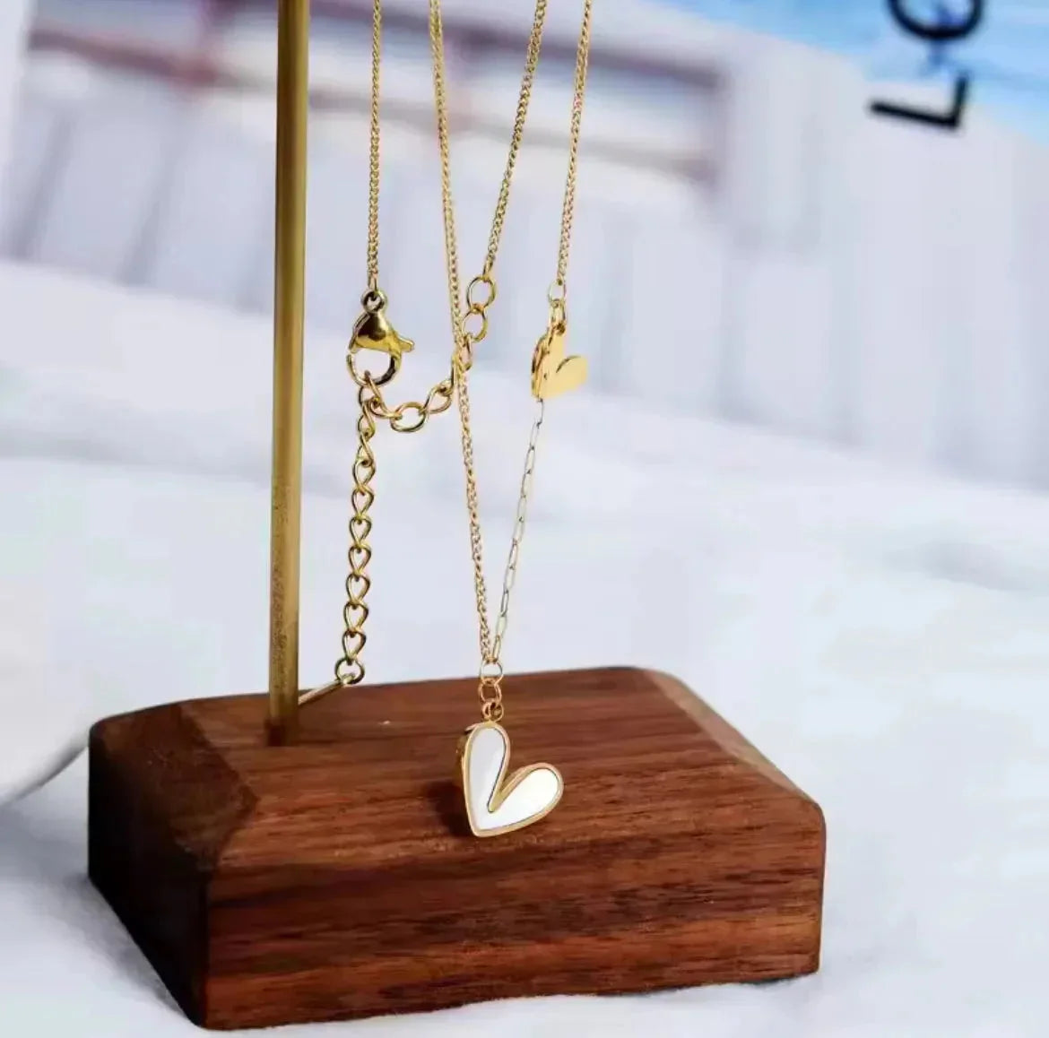 "Die goldene Halskette „Uschi“ von flyliving besteht aus einem weißen herzförmigen Anhänger mit Muschelfüllung und einem kleinen goldenen Charm, präsentiert auf einem Metallständer mit Holzsockel vor einem sanft verschwommenen Hintergrund."