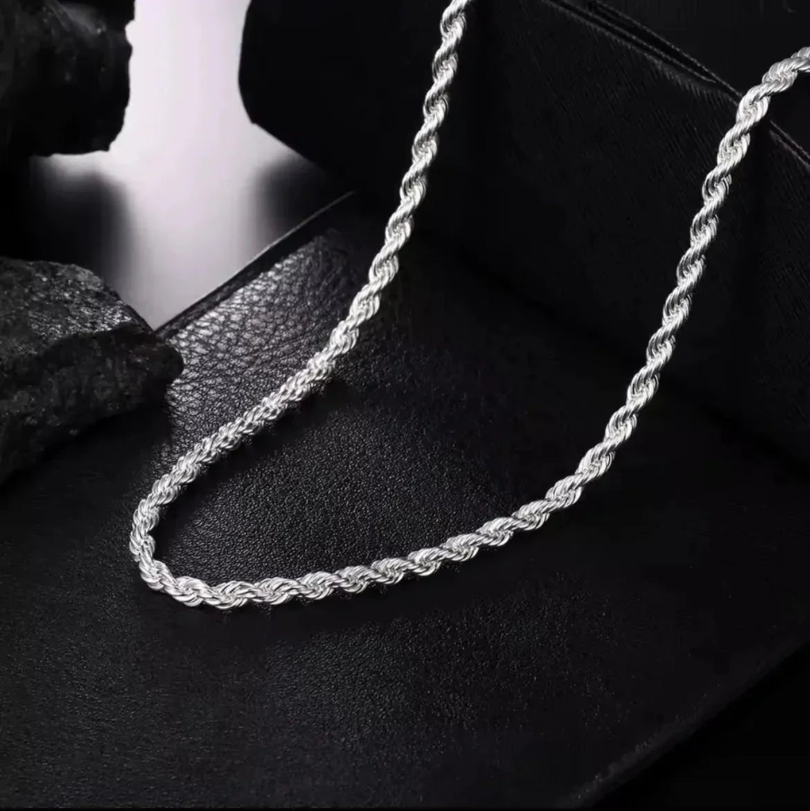 "Die Halskette „Helga“ aus Silber von flyliving mit filigran gedrehtem Design liegt auf schwarzem Leder, während dunkle Steine im Hintergrund die elegante Gliederkette stilvoll in Szene setzen."